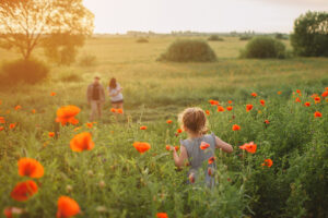 A young girl walking in a field of blooming poppies toward a smiling man and woman who are standing in a grassy field dotted with deciduous trees and shrubs.