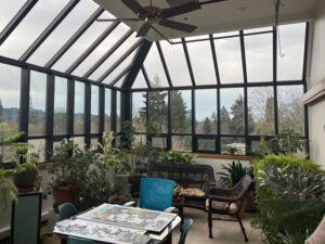 A tall sunroom with a ceiling fan, a wicker loveseat and chair, a table with a partially completed jigsaw puzzle, and numerous house plants of various sizes and types.